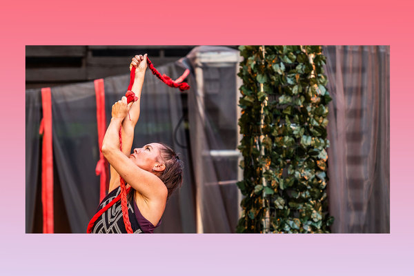 female dancer tightly gripping a long red rope with both hands, eyes looking up at it with fabric drapery backdrop.