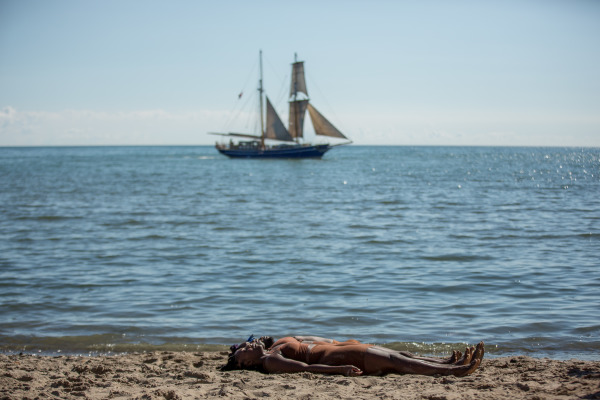 Two people laying on the sand at the beach with the water appearing behind them with a sail boat in the distance 