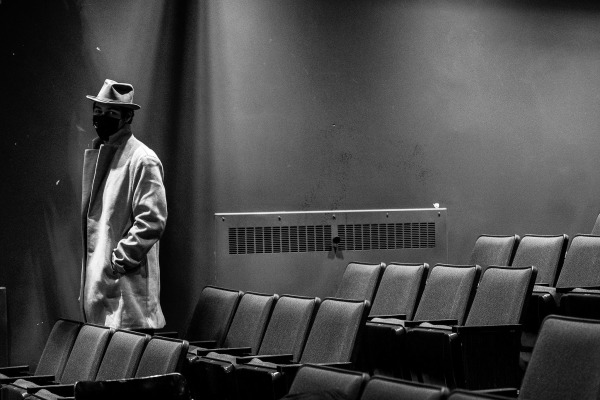 Male figure with face mask wearing hat and long trench coat in the auditorium seating. 