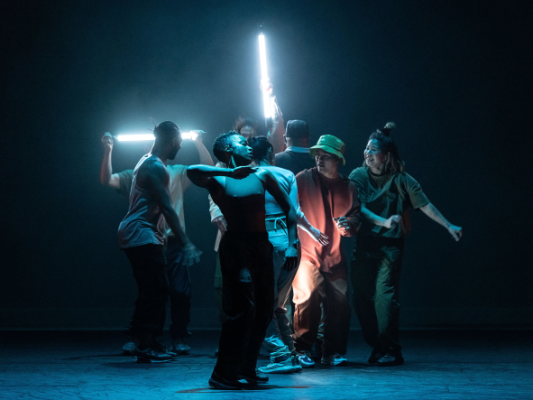 A group of dancers are illuminated from behind by bright neon lights.
