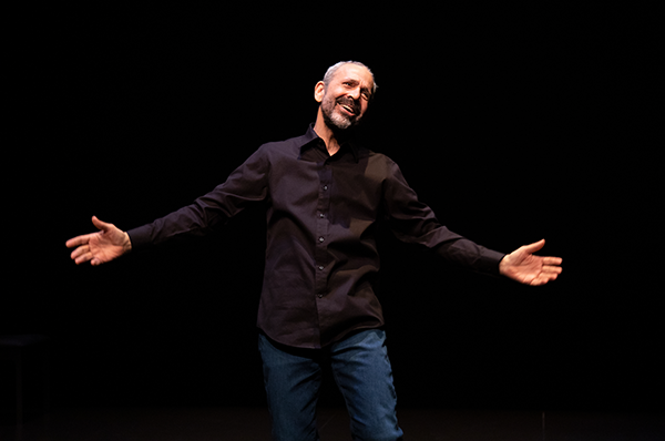 A white man in black button-up and jeans holds his arms open in welcome