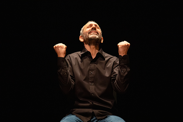 A man in a black button-up clenches his fists while looking toward the sky in longing