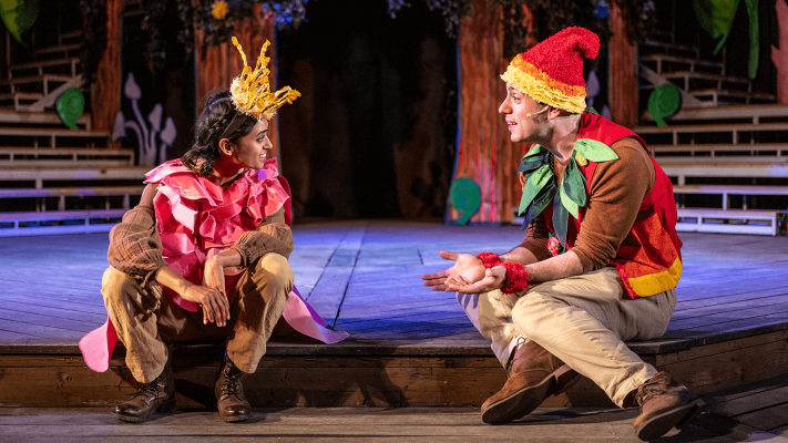Two actors dressed in bright reds and yellows sit at the lip of the stage talking with one another.