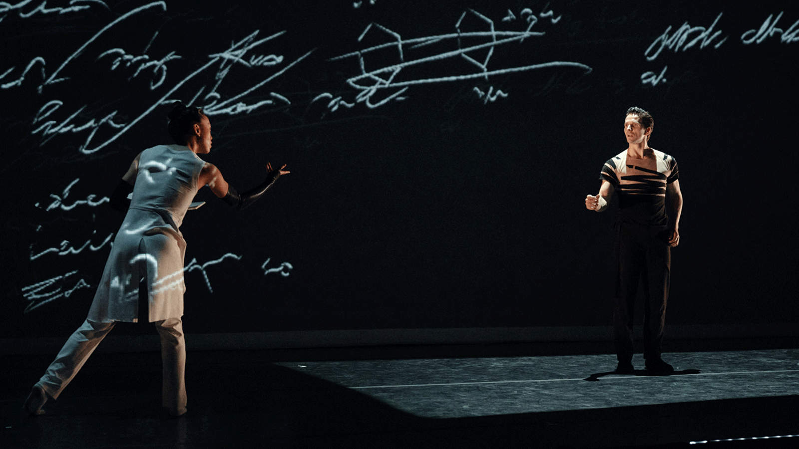 Two dancers reach toward each other across a stage with writing projected over them.