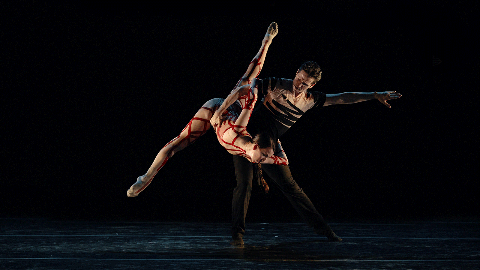 A male dancer lifts a female dancer to his side as she points her leg to the sky.