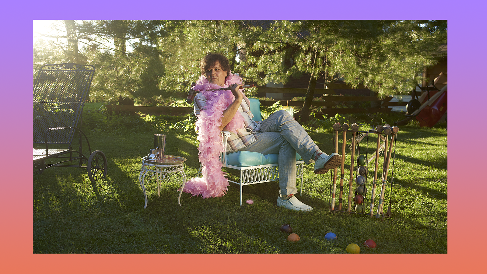 Ron Sexsmith wearing a feather boa sitting on a cushioned chair surrounded by trees