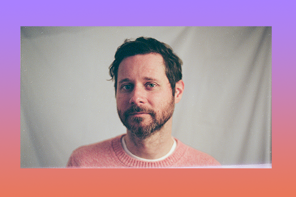 Dan Mangan, in a pink sweater, stands in front of a beige backdrop and looks into the camera.