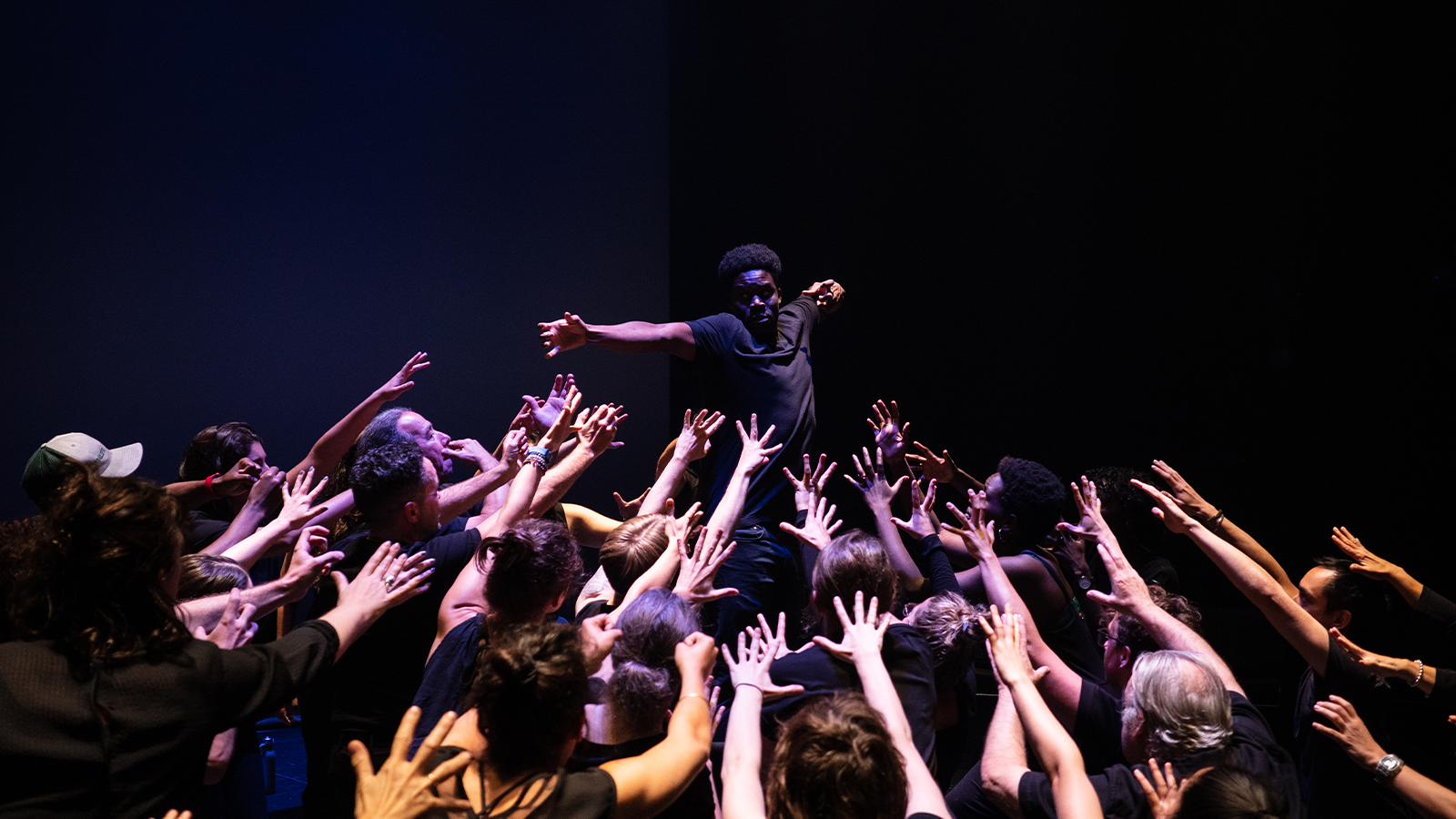 Dozens of outstretched hands reach out toward a Black performer obscured by shadow.